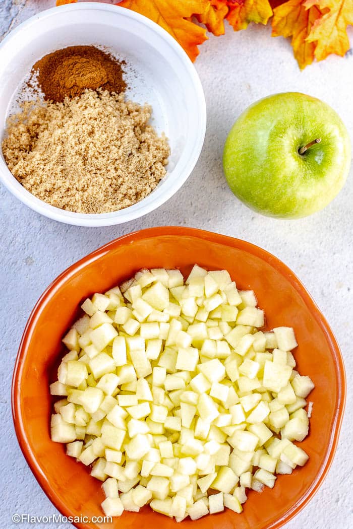 An orange bowl with chopped apples, a white bowl with brown sugar and cinnamon and a whole green apple on the side with a glimpse of fall leaves in the top right of the photo.