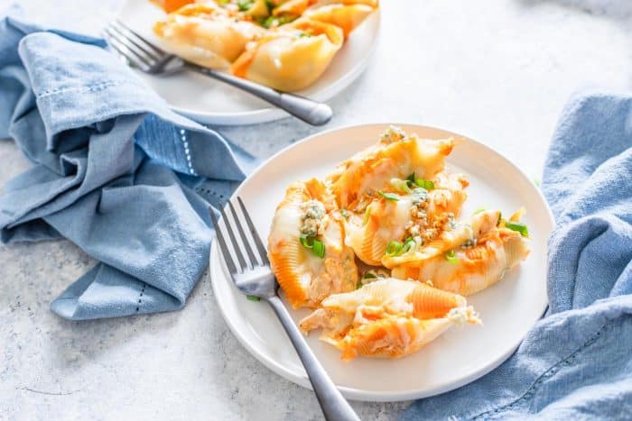 Horizontal photo of single serving of Buffalo Chicken Stuffed Shells on white plate with fork and blue napkins on side with a partial view of a second plate in the background.