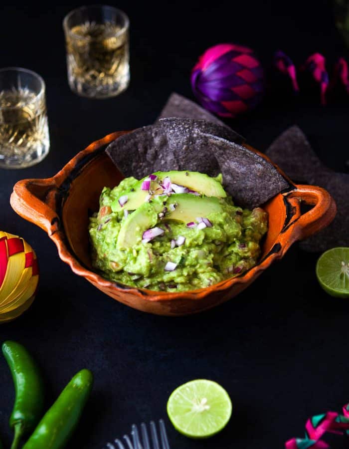 orange bowl with guacamole with dark tortilla chips with a black background surrounded by shot glasses, lime slices, chilies and a pink and purple decoration.