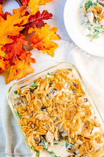 Overhead view of cooked green bean casserole with one spoonful removed and a white plate with a partial view of a single serving in the upper right corner with fall leaves as decoration in the upper left corner.