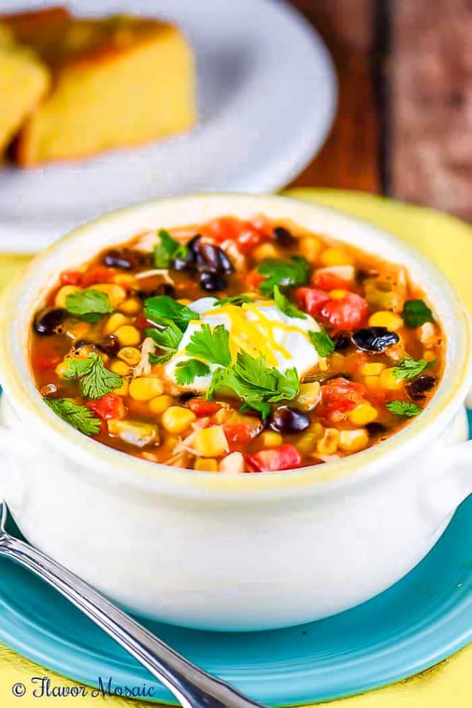 View of white soup bowl with Slow Cooker Enchilada Soup with black beans, corn, tomatoes, cilantro, cheese, and sour cream.