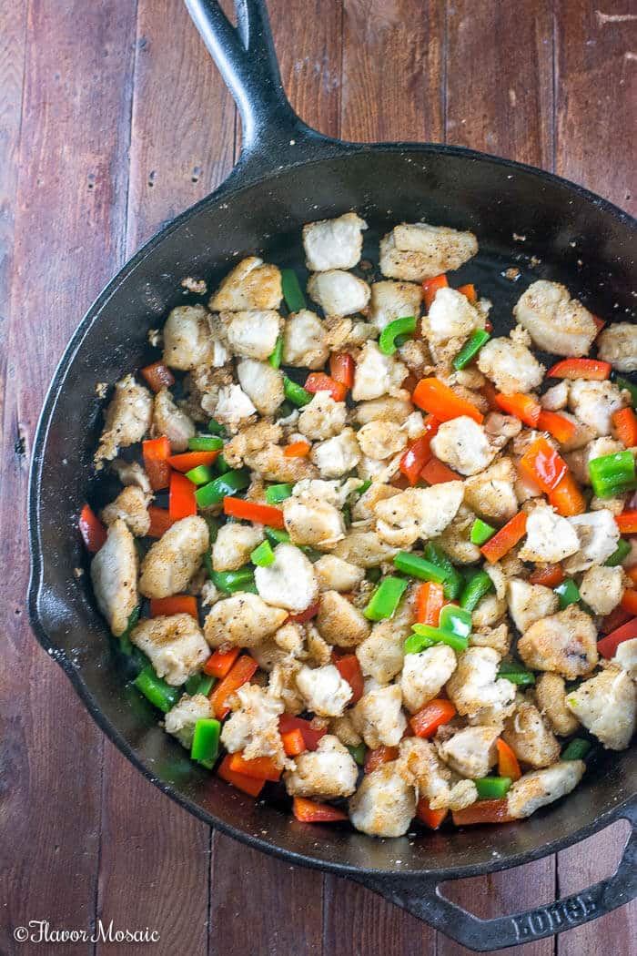 Overhead photo of chicken cubes covered in cornstarch being cooked in a cast iron skillet with red and green bell peppers.