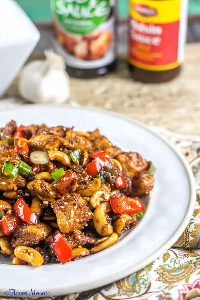 A Easy Spicy Chinese Cashew Chicken serving on a white plate with jars of soy sauce and hoisin sauce, along with some garlic in the background.
