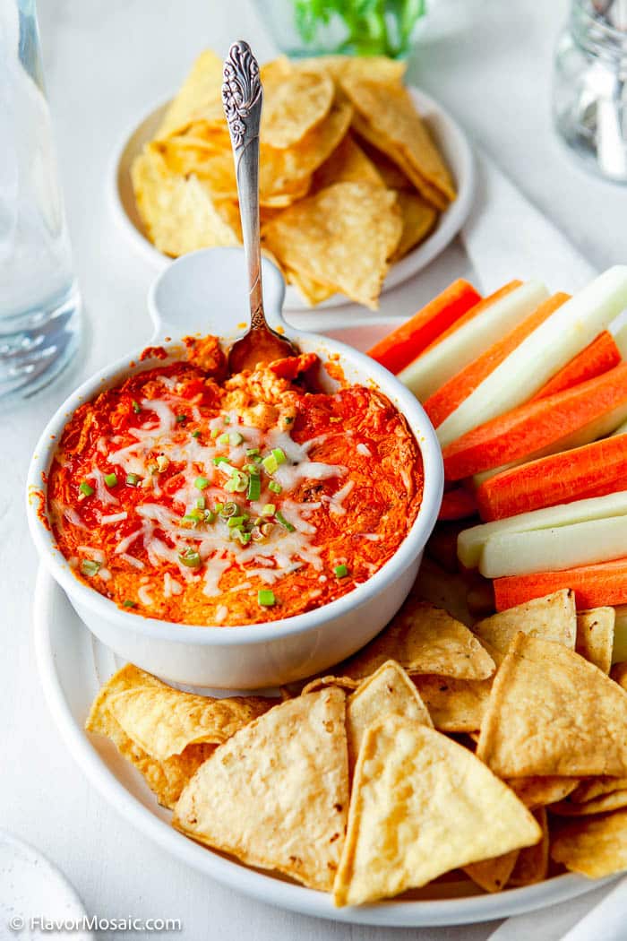 white bowl with Buffalo Chicken Dip topped with white cheese and green onions, surrounded by carrots and tortilla chips with a white background.