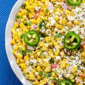 Overhead view of Mexican Street Corn Salad, topped with sliced jalapenos, crumbled cotija cheese, and chopped fresh cilantro, in a white bowl with a blue background.