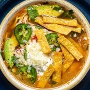 Overhead photo of a white bowl of Chicken tortilla Soup topped with tortilla strips, sliced jalapeños, and cilantro, sitting on a blue plate.