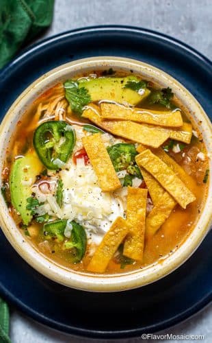 Overhead photo of a white bowl of Chicken tortilla Soup topped with tortilla strips, sliced jalapeños, and cilantro, sitting on a blue plate.