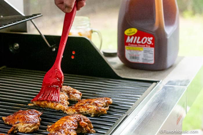 Brushing Sweet Tea BBQ Sauce on Chicken thighs on grill with a jug of sweet tea in the background.