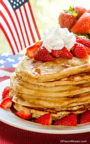 Stack of Strawberry Pancakes topped with strawberries, maple syrup, and whipped cream, on white plate with bowl of strawberries in the background.