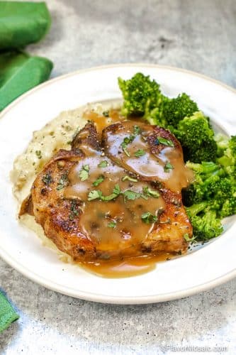 Vertical photo of Instant Smothered Pork Chops sitting on white plate with sides of broccoli and mashed potatoes.