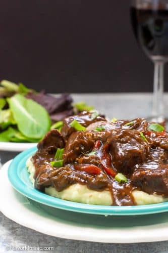 An individual serving of Instant Pot Beef Tips and Gravy served over mashed potatoes on a blue plate sitting on a larger white plate with a salad and glass of wine in the background.