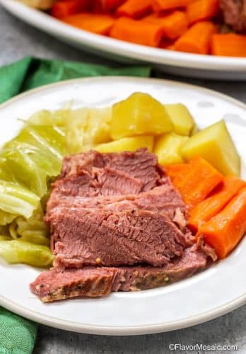 White plate with serving of corned beef with cabbage, carrots, and potatoes, with green napkin on the side and a partial view of serving platter in the background.