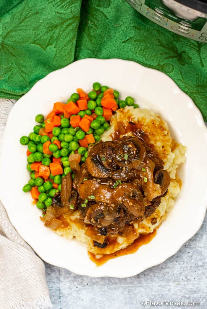Overhead photo of white plate with Salisbury Steak with Onion and Mushroom Gravy with mashed potatoes and peas and carrots.