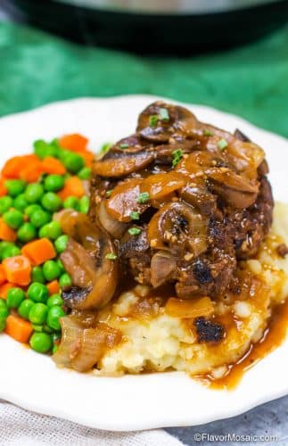 White plate with Instant Pot Salisbury steak with gravy, onions and mushrooms on top of mashed potatoes and served with peas and carrots.