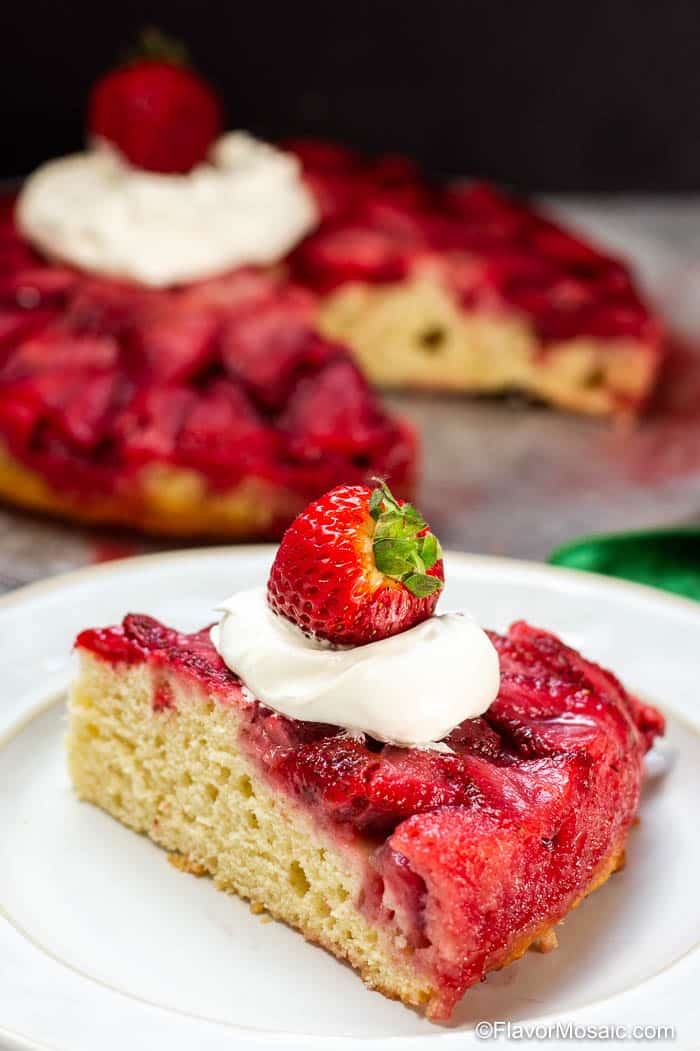 Single serving serving Strawberry Upside Down Cake on white plate topped with whipped cream and whole strawberry with whole cake in the background.