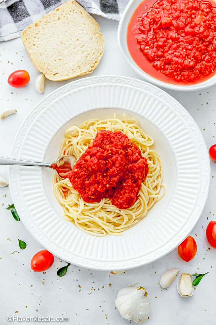 overhead view of white bowl with spaghetti topped with marinara sauce surrounded by cherry tomatoes and fresh garlic and basil with a bowl of marinara sauce and a slice of bread in the background.