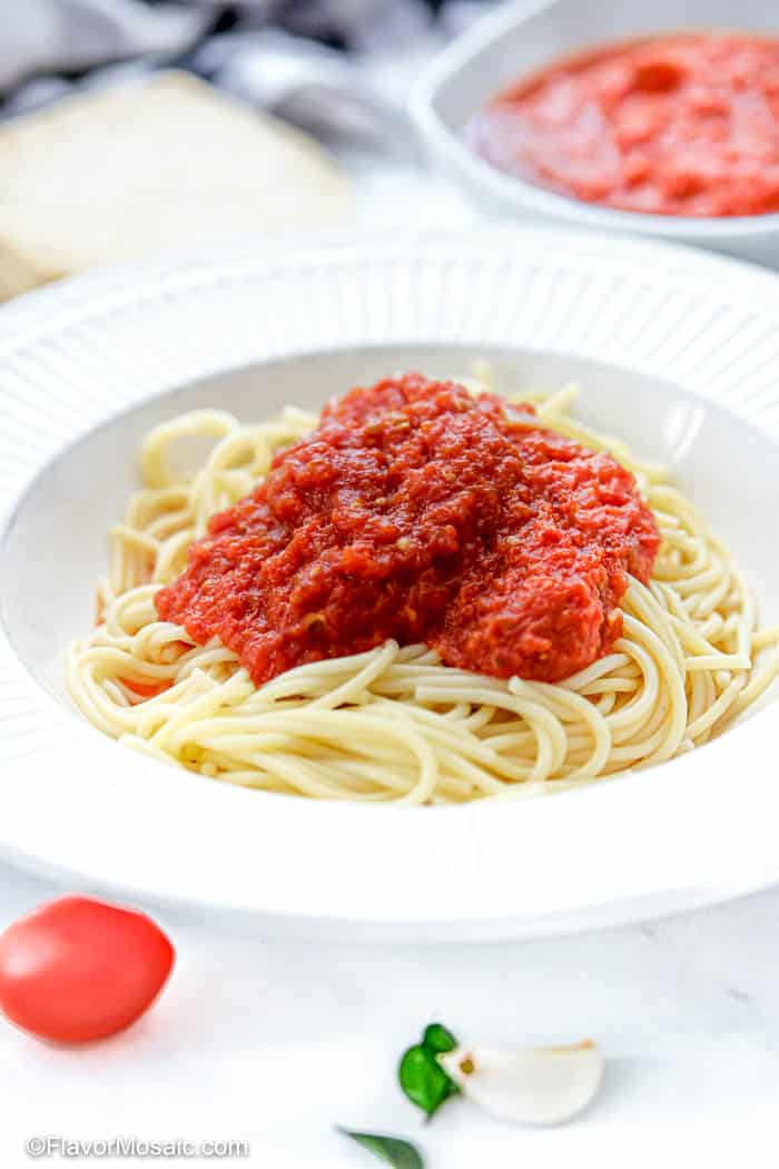 White bowl of spaghetti topped with marinara sauce with Roma tomato and garlic in the foreground and a small bowl of marinara sauce and bread in the background.