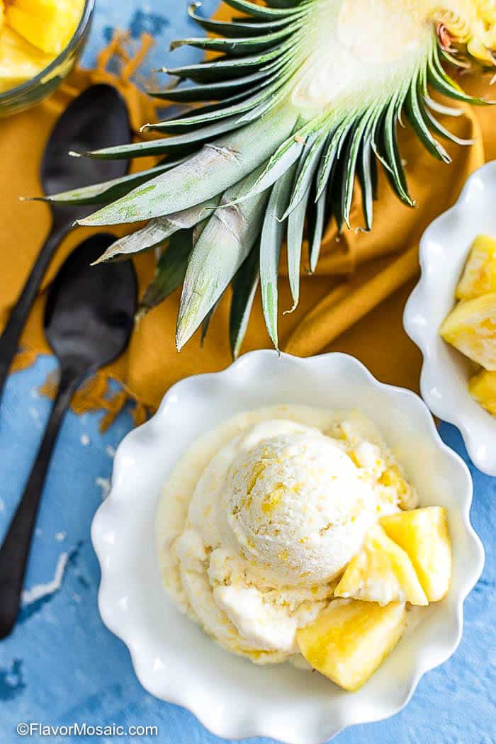 Overhead view of single serving of pineapple ice cream in white bowl with blue background with whole pineapple sliced vertically with orange napkin and 2 spoons.