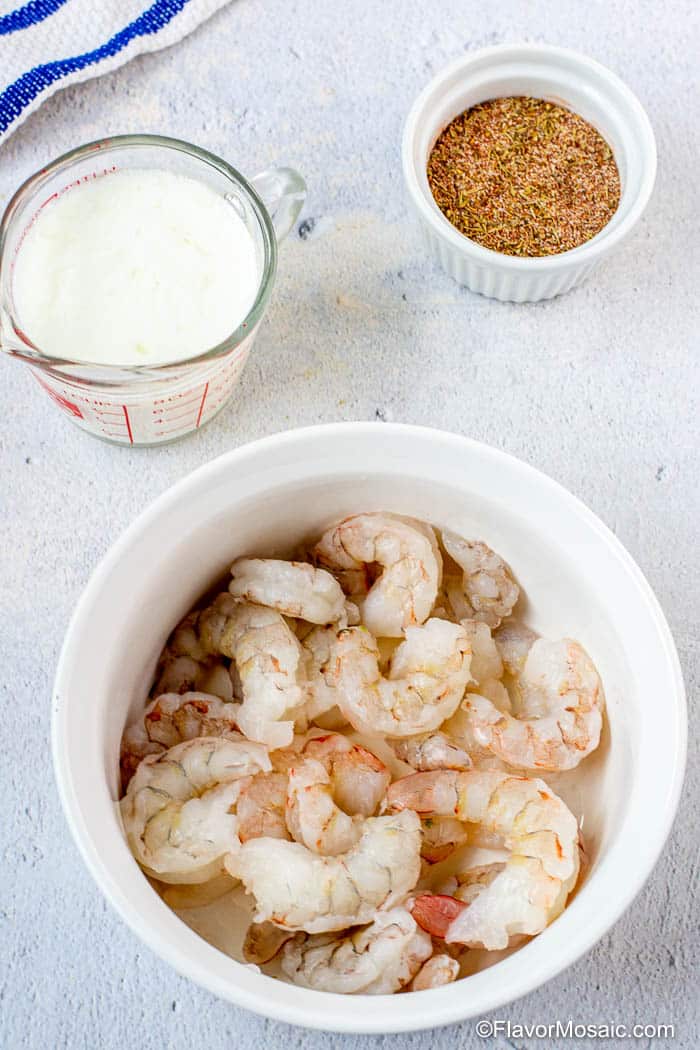 White bowl of raw shrimp with measuring cup of buttermilk and a small bowl of blackened seasoning in the background.