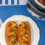 Overhead view of 2 dressed hot dogs on white plate with blue background with partial view of Instant Pot with hot dogs.