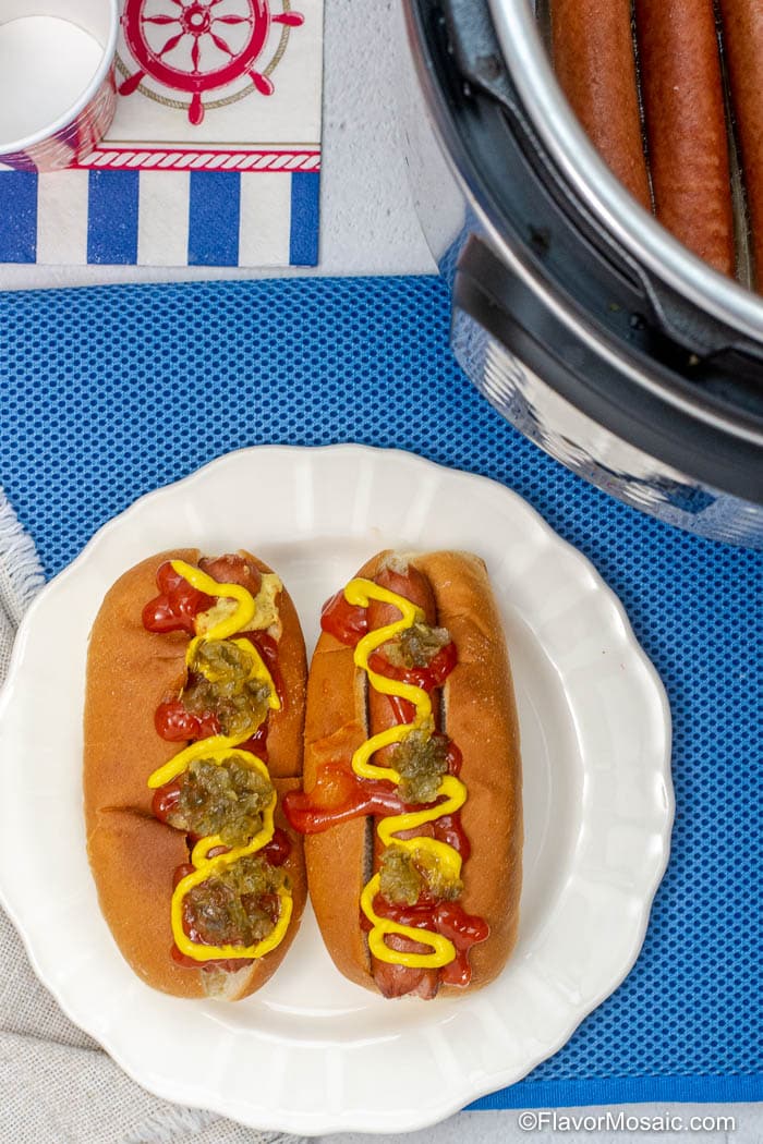 Overhead view of 2 dressed hot dogs on white plate with blue background with partial view of Instant Pot with hot dogs.