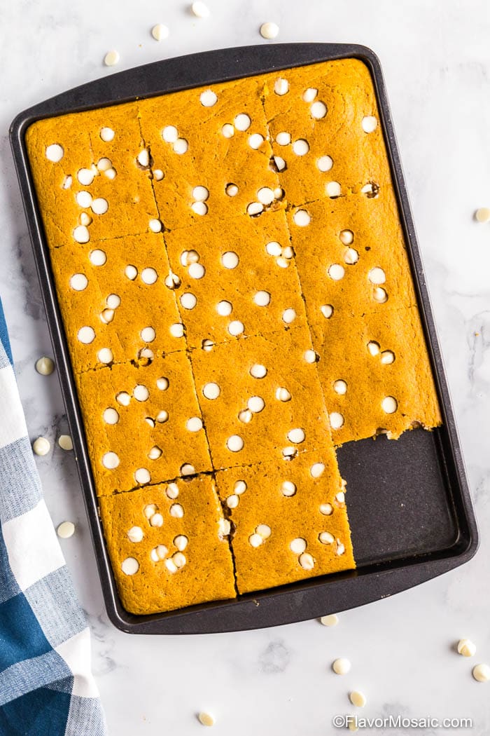 Overhead view Pumpkin Sheet Pan Pancakes in baking pan with blue and white checkered napkin on the left side and white chocolate chips scattered around it on a marble counter.