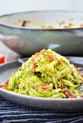 Plate with a heaping pile of zucchini noodles with pesto and bacon with a large bowl in the background.