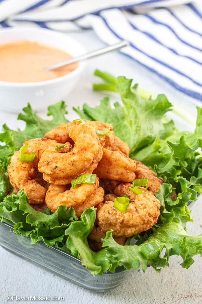 Photo of Bang Bang Shrimp on top of green leaf lettuce topped with green onions with a bowl of bang bang sauce in the background and a white napkin with blue stripes in the top right corner.