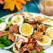 Spinach Salad with Warm Bacon Dressing on white plate with hard boiled eggs, bacon, sliced onions, and sliced mushrooms, with fall colored leaves on blue fabric and a bottle of bacon dressing in the background.