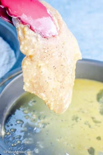 View of egg covered chicken breast held in the air with tongs above a pan with the egg mixture for breading the chicken breast.
