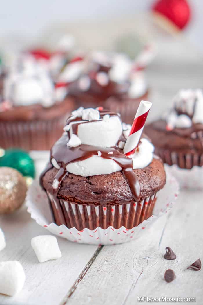 Single Hot Chocolate Cupcake topped with marshmallow and chocolate ganache with cupcakes in the background and green and gold Christmas ornaments on the left side.
