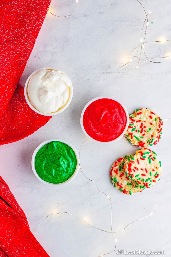 Overhead view of three bowls of buttercream icing in 3 different colors, red, green, and white, with 3 sprinkled-covered thumbprint cookies next to them.