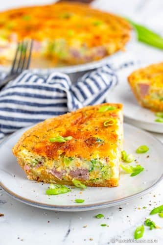 View on a single slice of ham and broccoli quiche on a white plate with a fork and blue and white striped napkin, as well as a whole quiche and partial view of the corner of another slice of quiche in the background.