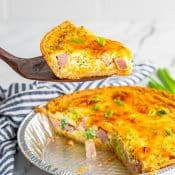 Spatula holding up 1 slice of Ham Broccoli and Cheese Quiche over the entire pie below with a blue and white napkin in the back.