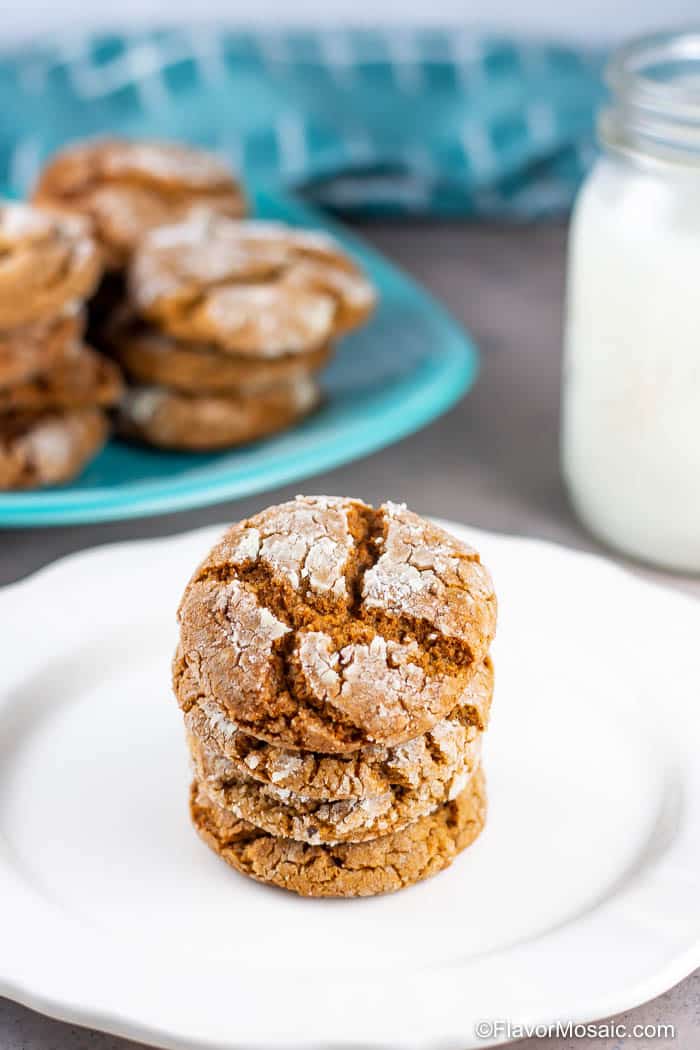 4 molasses crinkle cookies stacked high on a white place with a platter of cookies and a glass of milk in the background.