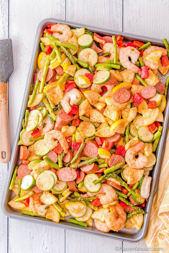 Overhead view of Cajun shrimp, Kielbasa sausage, and vegetables on sheet pan with white wood background.