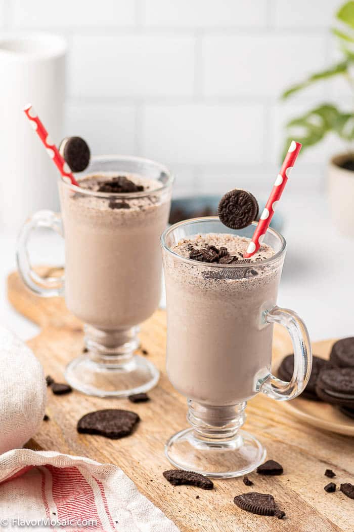 Two Tall Glasses with Oreo Milkshakes topped with Oreo pieces and surrounded by Oreo cookie pieces on a cutting board
