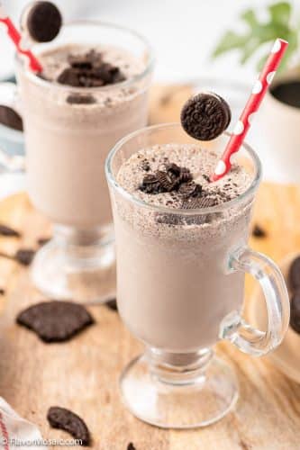 Two Tall Glasses with Oreo Milkshakes topped with Oreo pieces and surrounded by Oreo cookie pieces on a cutting board