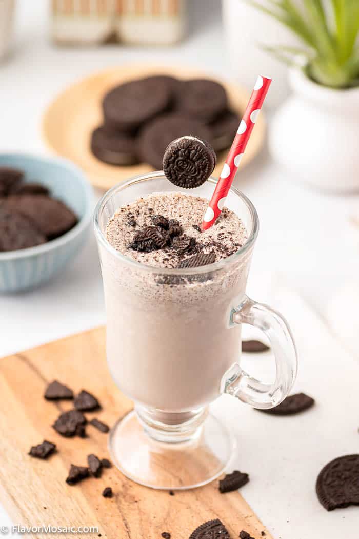 Tall Glass with an Oreo Milkshake topped with Oreo pieces and surrounded by Oreo cookie pies on a cutting board with a bowl and a plate of Oreos and a plant in the background 