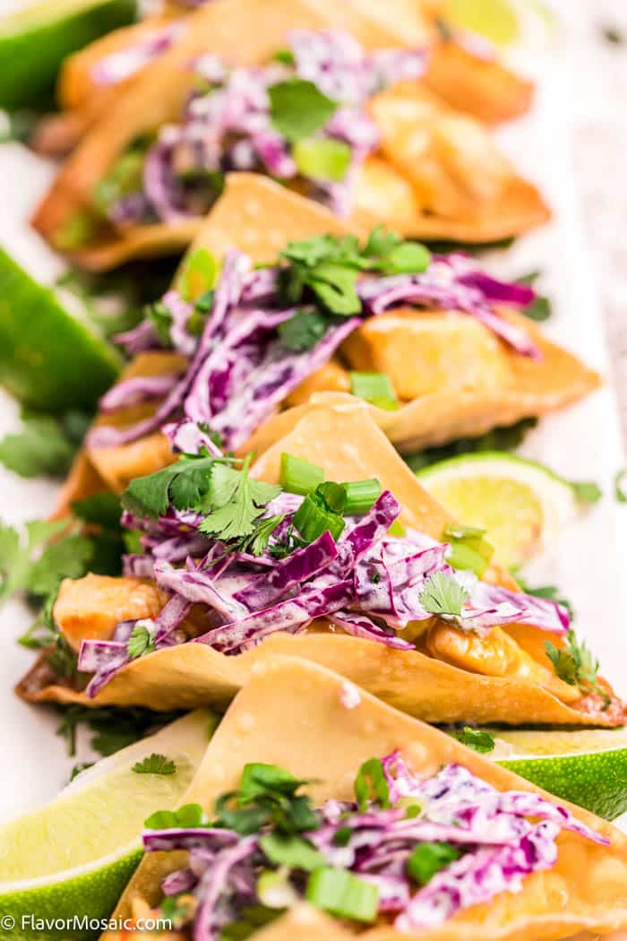 Several Chicken Wonton Tacos lined up vertically served with red cabbage slaw, cilantro and limes.