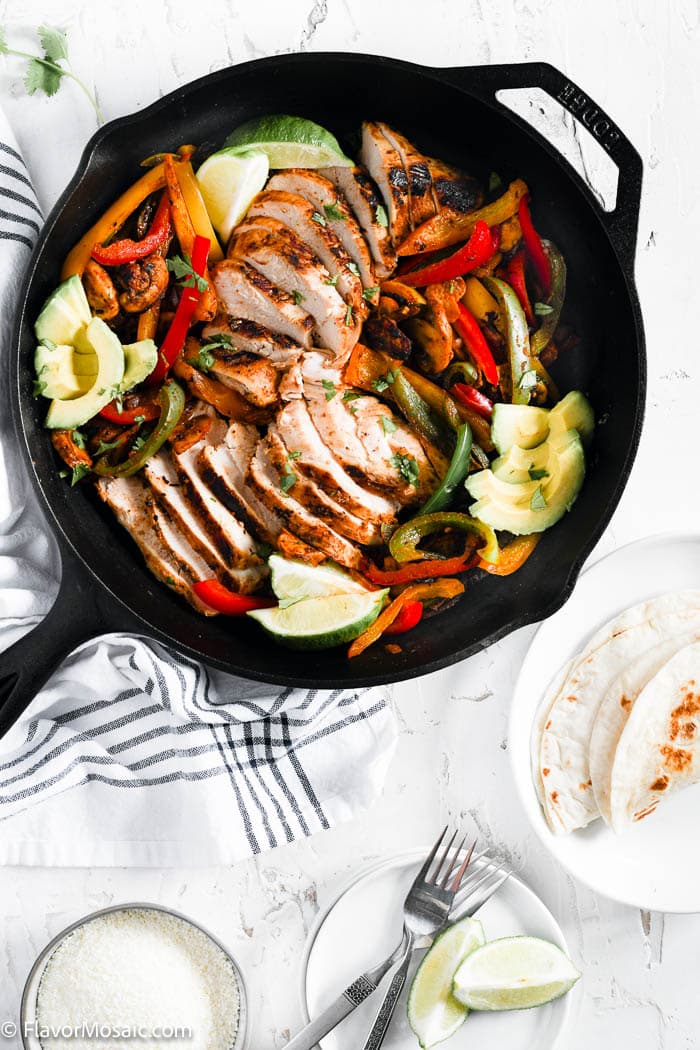 Overhead view of chicken fajitas in cast iron skillet on white background with a napkin, forks, limes, and tortillas on the side.
