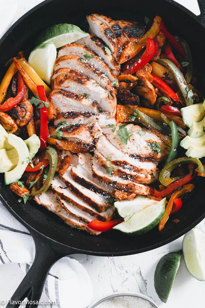 Overhead view of sliced skillet chicken fajitas in a cast iron skillet with sliced bell peppers, avocados, limes, and onions.