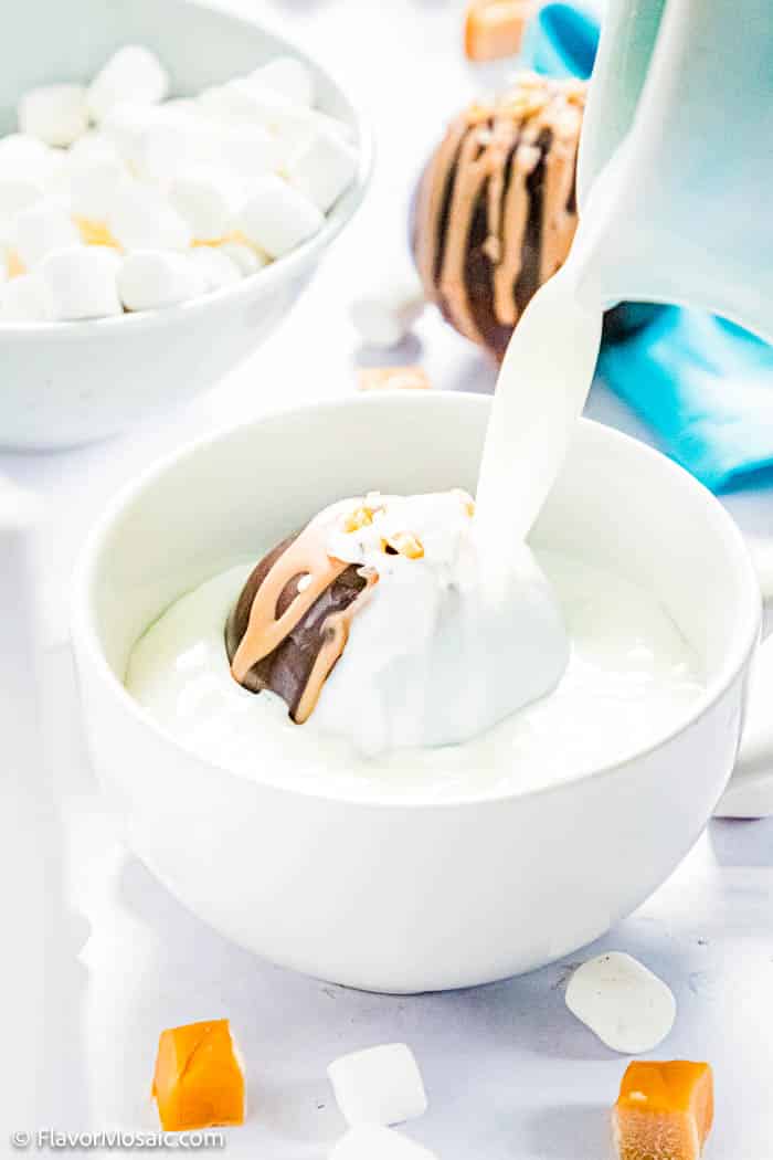 Pouring milk over a salted caramel hot chocolate bomb in a white bowl surrounded by caramel pieces and mini marshmallows with a white bowl of mini marshmallows in the left corner with a hot chocolate bomb next to it and a blue napkin in the top right corner.