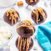 2 hot chocolate bombs drizzled with salted caramel stacked on top surrounded by caramels, marshmallows and a blue napkin with 3 salted caramel hot chocolate bombs in the background.