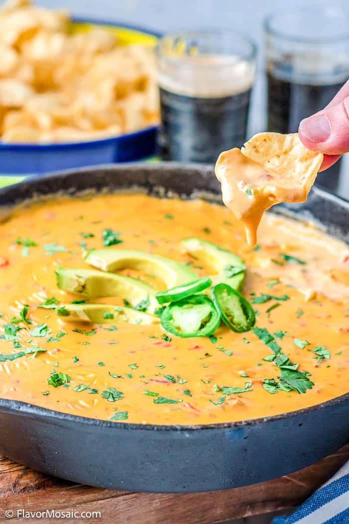 Skillet of loaded queso topped with sliced avocados,, jalapenos, and chopped cilantros, with a tortilla chip held over the skillet of queso with queso dripping off the chip into the skillet.