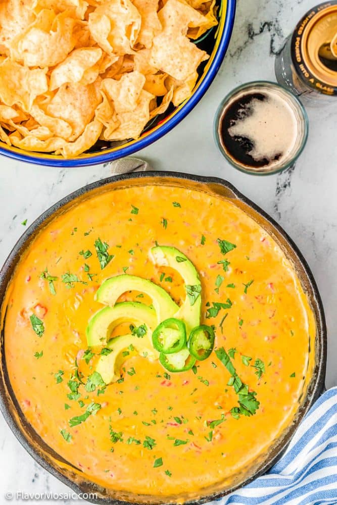 Overhead view of large cast iron skillet of queso dip topped with sliced avocado and chopped cilantro, next to a bowl of tortilla chips and glass of beer wuth a beer can next to it and a blue and white striped napkin in the bottom right corner.