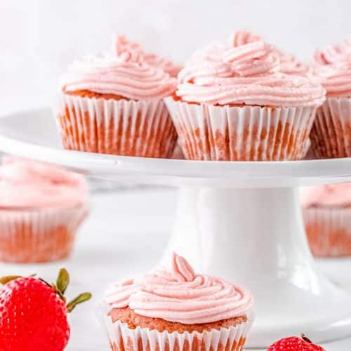Side view of a cake stand with strawberry cupcakes with a cupcake under it surrounded by whole strawberries and more strawberry cupcakes in the background.