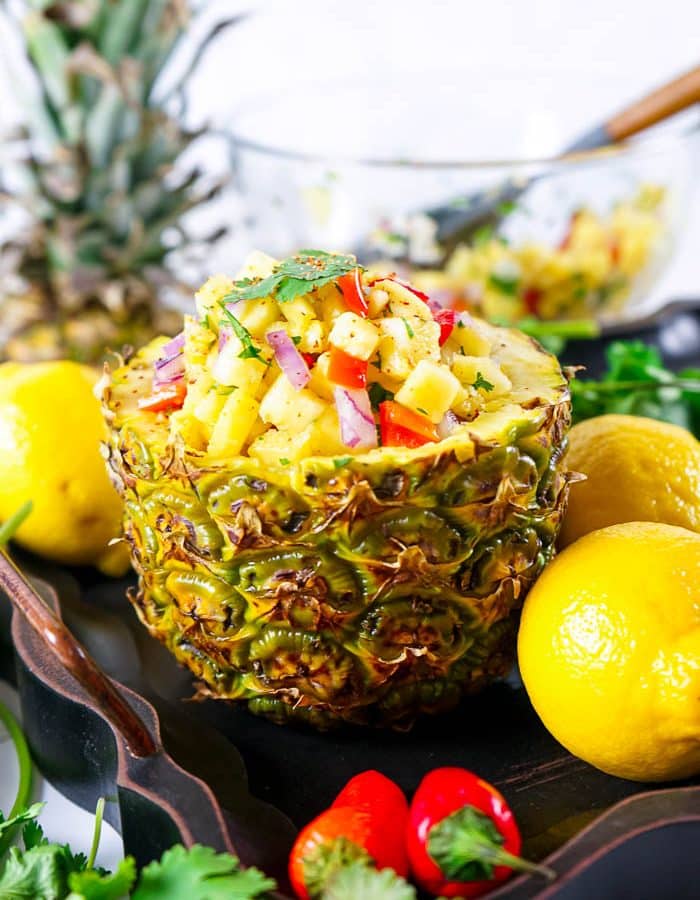 Side view of bottom half of pineapple, hollowed out and filled with pineapple salsa, surrounded by 3 whole lemons, 2 red chili peppers, cilantro, the top half of the pineapple in the back, along with a glass bowl of the remaining salsa.