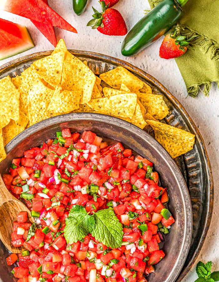Overhead view of bowl with watermelon salsa surrounded by tortilla chips and slices of watermelon, whole jalapeños, strawberries, and a green napkin in the top right.