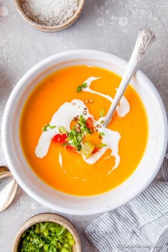 Overhead view of a small white bowl with a single serving of Instant Pot Butternut Squash Red Pepper Soup garnished with swirls of sour cream, chopped green onions, and chopped red bell peppers.
